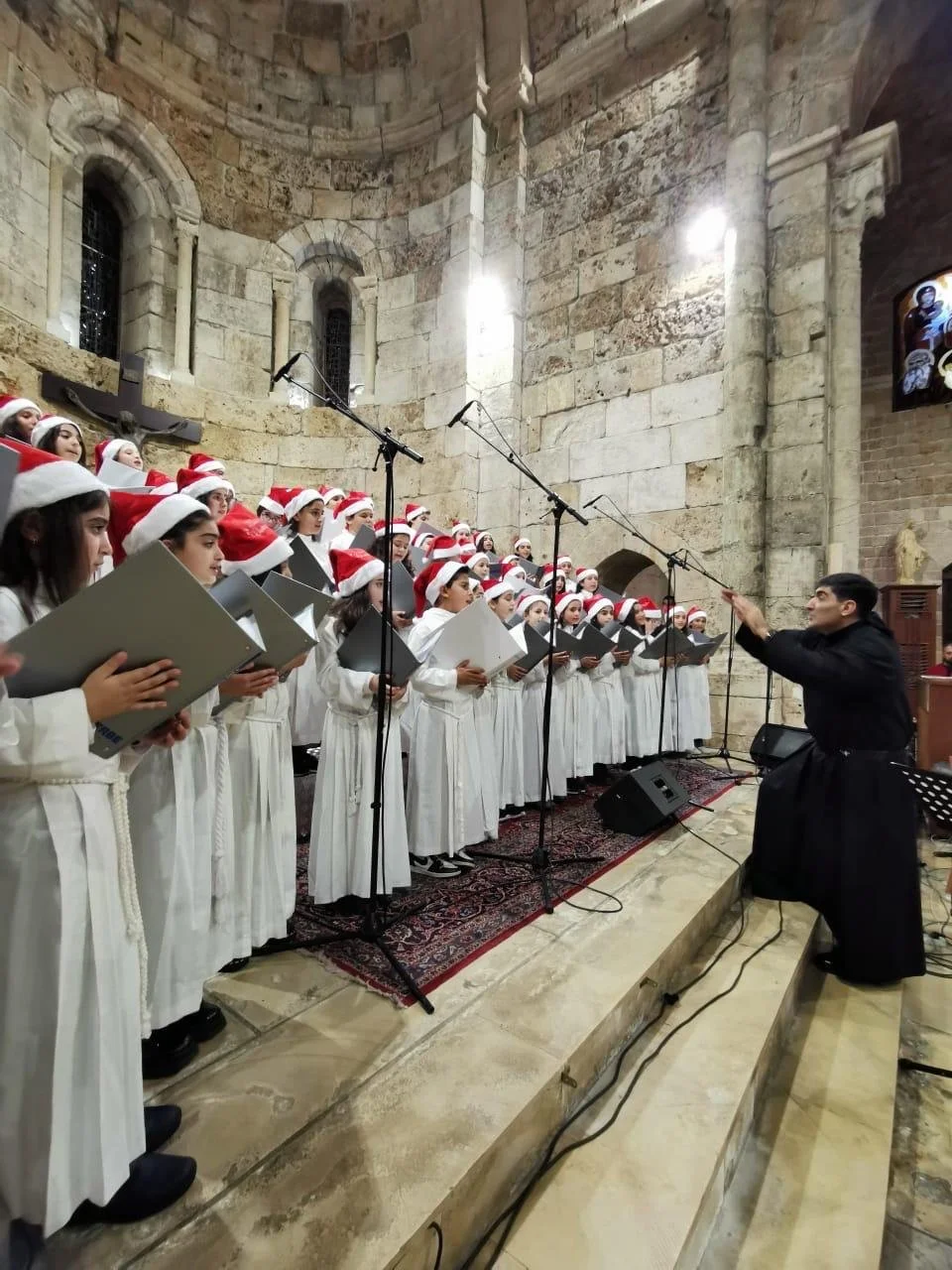 A children's choir wearing white robes and Santa hats singing on stage, led by a conductor in black robes, inside a historic stone church with small arched windows.