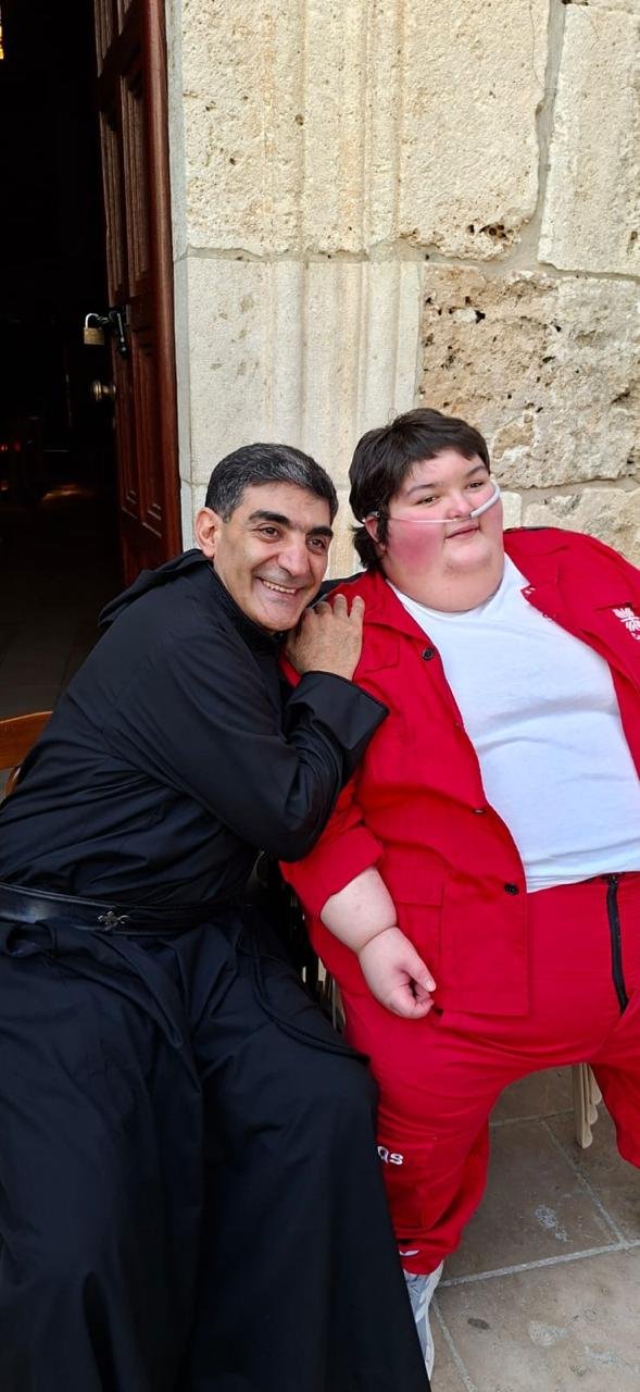 A man with dark hair, smiling, wearing a black jacket, sits next to a large boy with a nasal tube, wearing a red jacket and white shirt, in front of a stone wall.