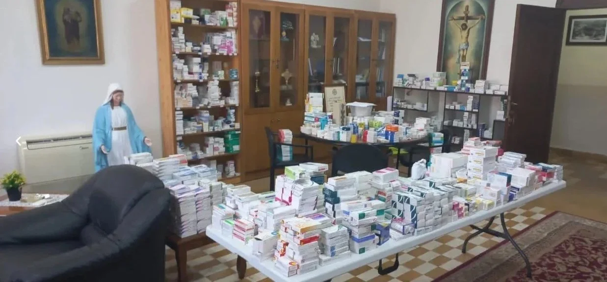 Table filled with numerous stacks of medications, with cabinets of more medication behind, in a room with religious decorations.