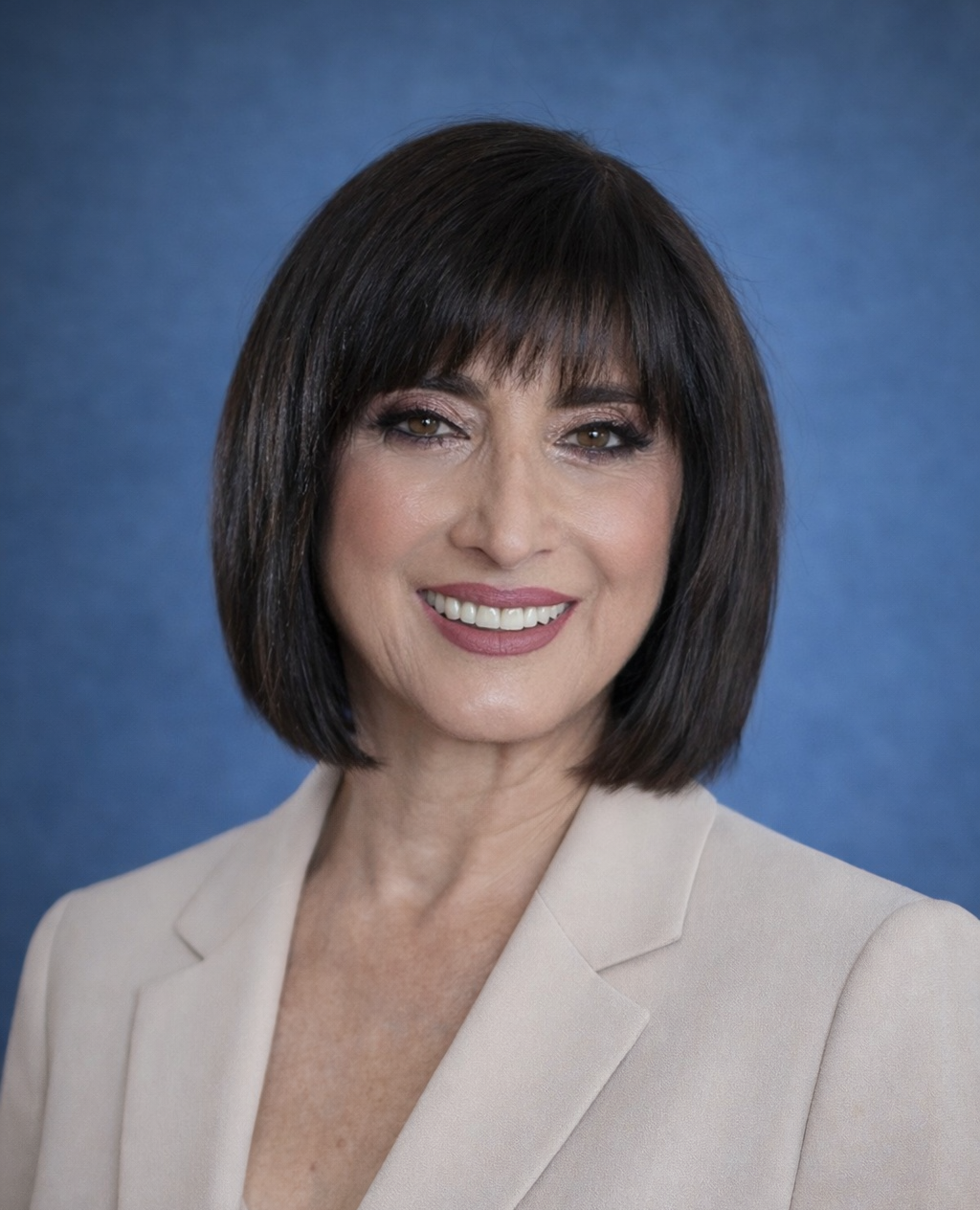 A professional woman with shoulder-length dark brown hair, wearing a light-colored blazer, smiling against a blue background.