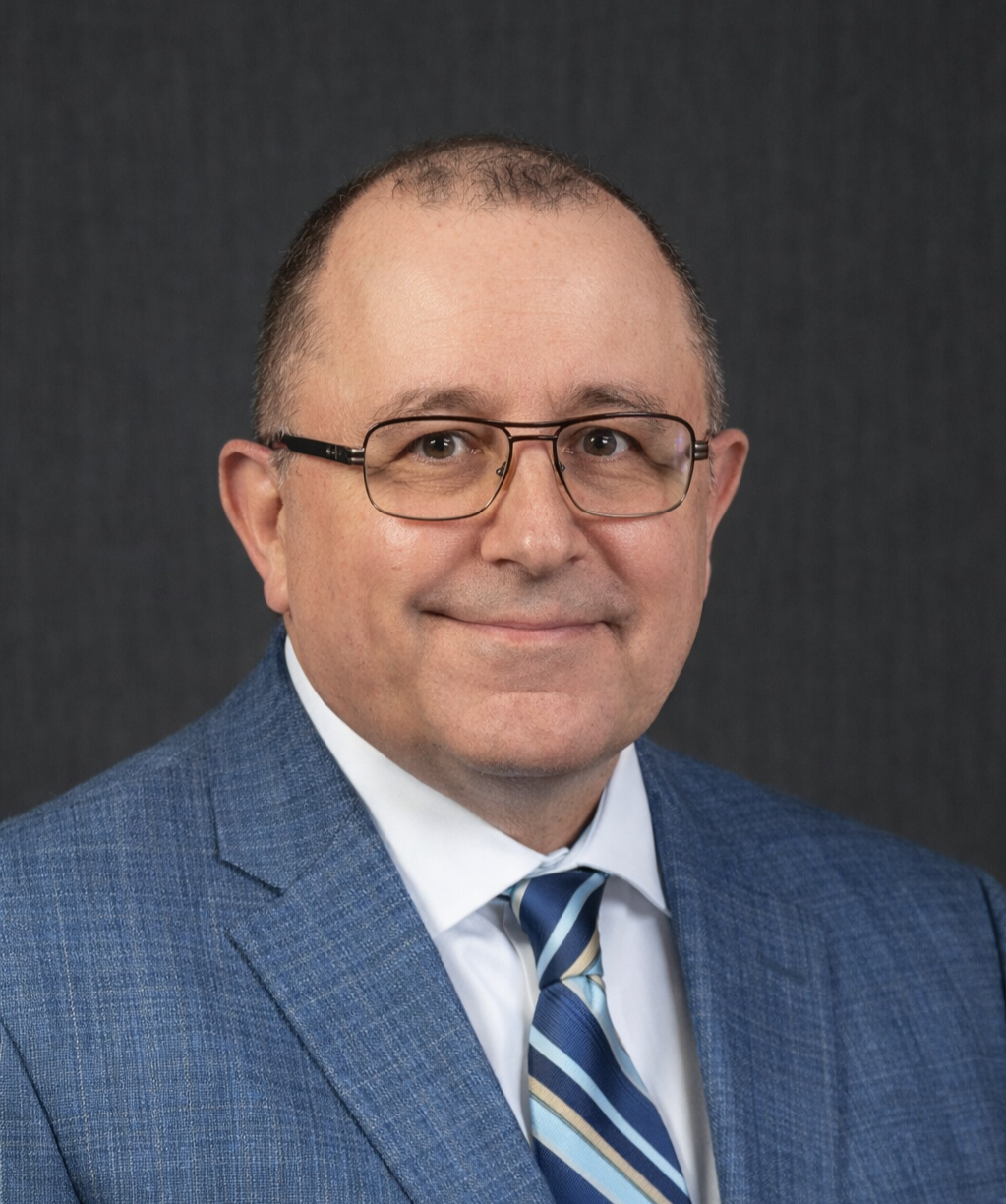 A middle-aged man with glasses smiling, wearing a blue suit, white shirt, and a striped blue tie, against a dark background.