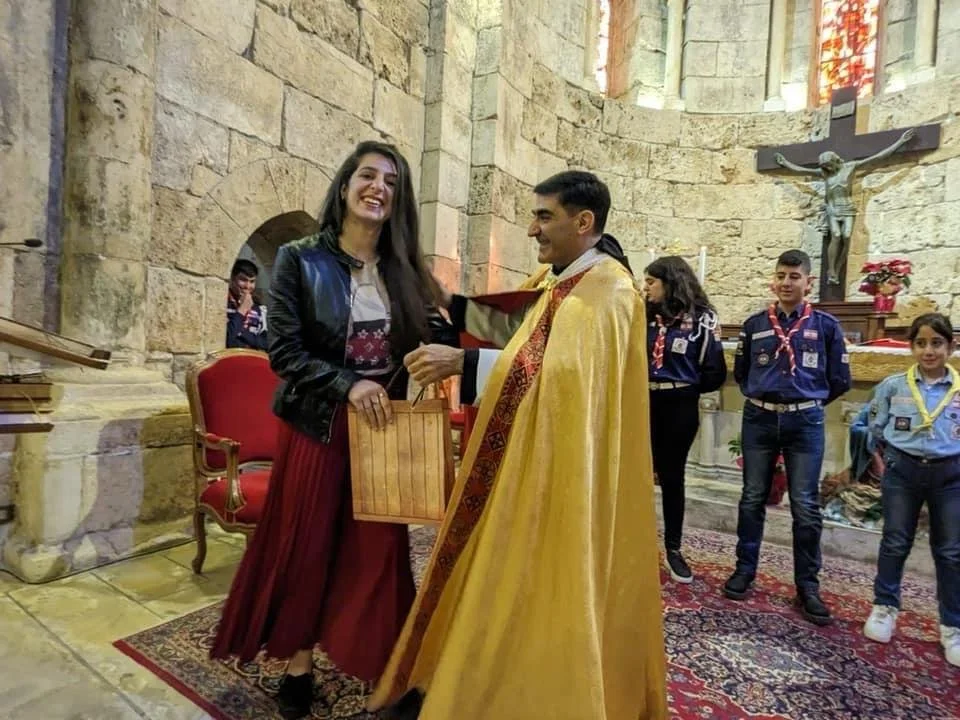 A woman receiving a gift from a priest inside a church, with four scouts standing in the background. The woman is smiling, wearing a black jacket and a long red skirt. The priest is wearing ceremonial robes and a gold cape, and is handing her a gift bag. The church has stone walls, an altar with a crucifix, and a decorative rug on the floor.