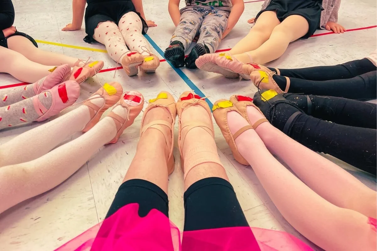 Children sitting in a circle at a ballet class, wearing pointe shoes and tights.