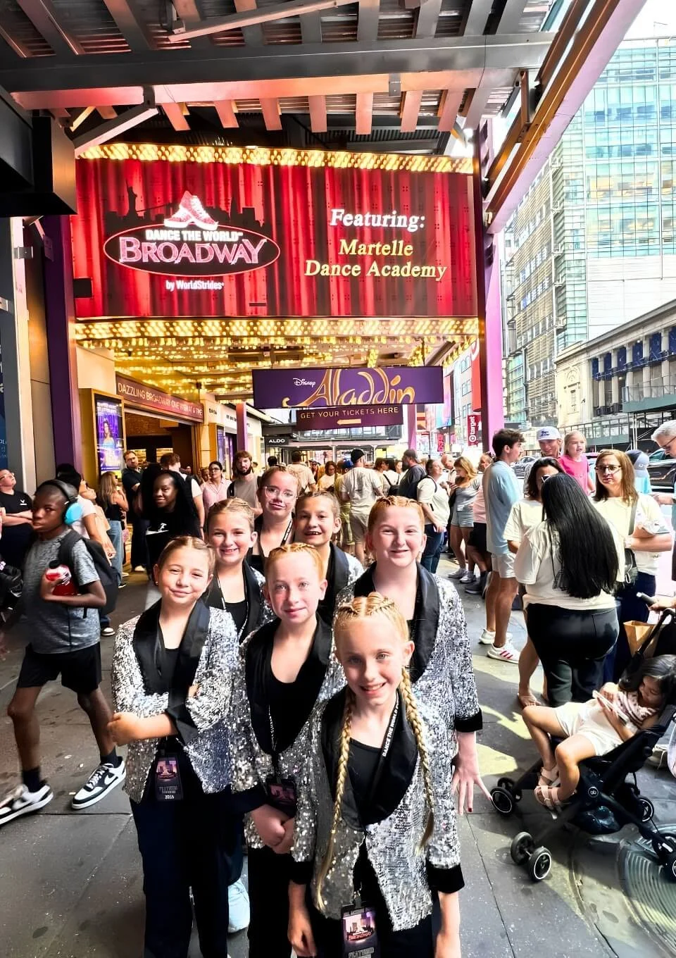 Group of young girls in coordinating outfits posing in front of a Broadway theater in New York City, on a busy city street with bright theater marquee and crowds.