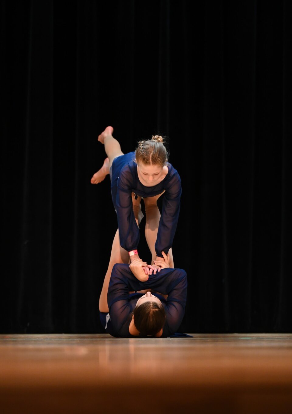 Two women performing a synchronized acrobatic yoga pose on a stage with a black curtain background.