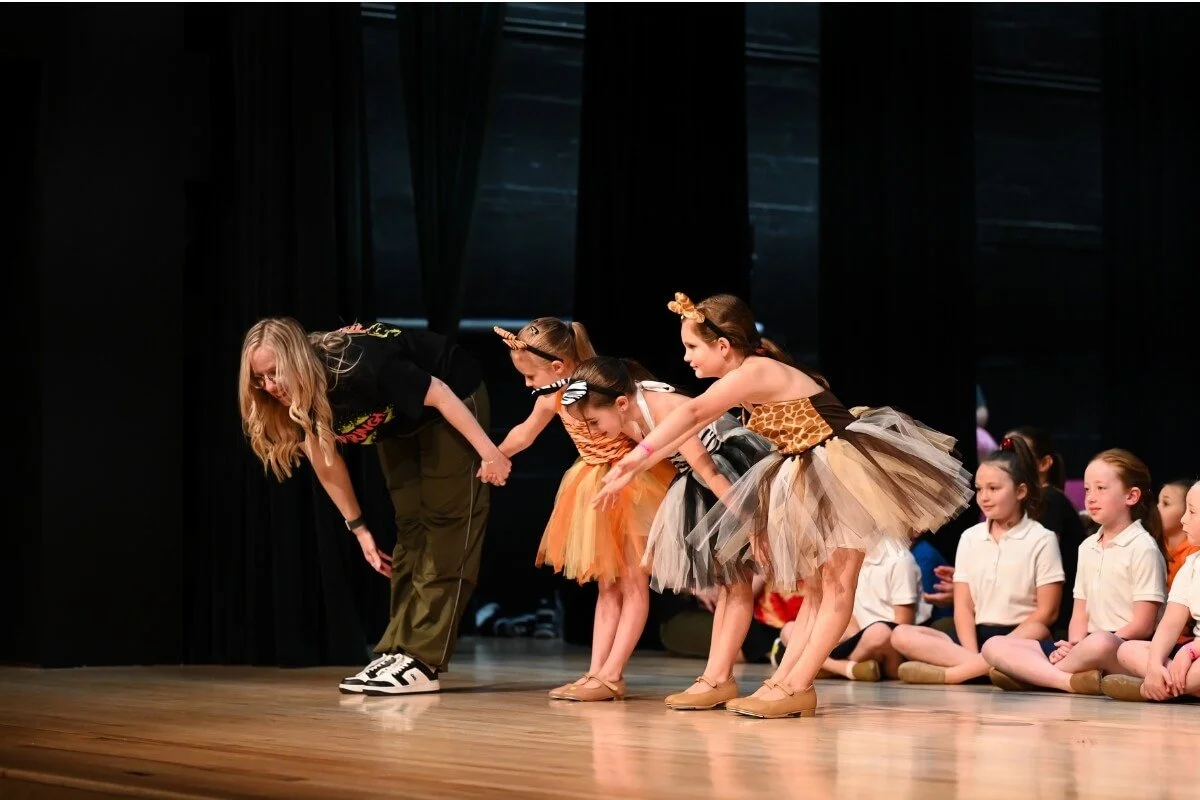 A dance teacher with long blonde hair bowing at the front of a stage while four young girls dressed as tigers bow in response, with other children sitting on the stage behind them.