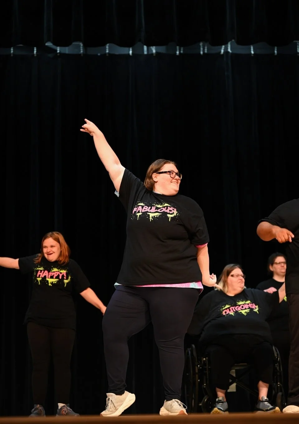 Group of diverse women dancing on stage, wearing black t-shirts with colorful words. They are smiling and having fun against a black curtain background.