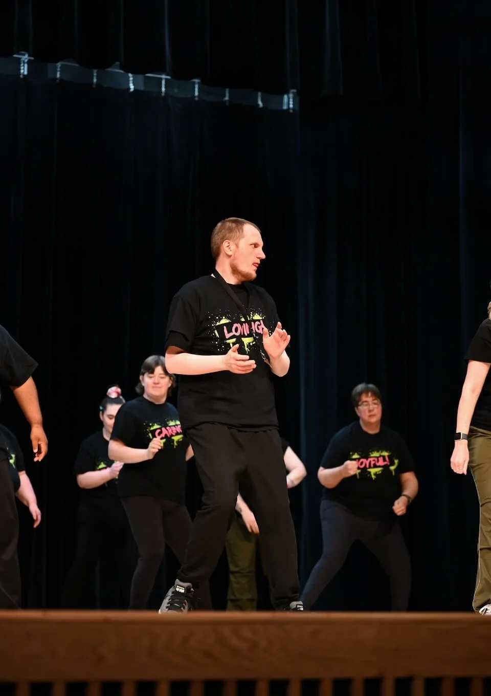 Group of people on stage practicing a dance, wearing black t-shirts with colorful text, and a black curtain in the background.