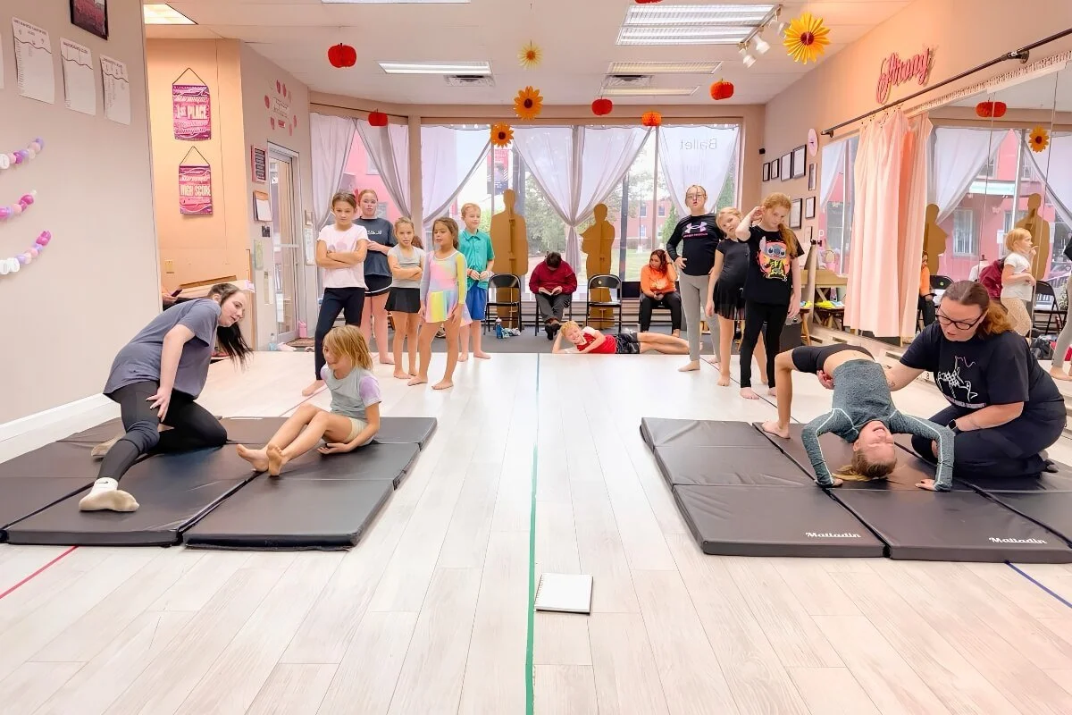 Children and instructors in an acro class in a bright studio with pink and yellow decorations, practicing ground exercises and watching others.