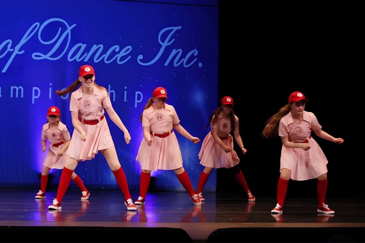 Five young girls dancing on stage in matching pink uniforms with red hats, red socks, and sneakers, performing at a dance competition.