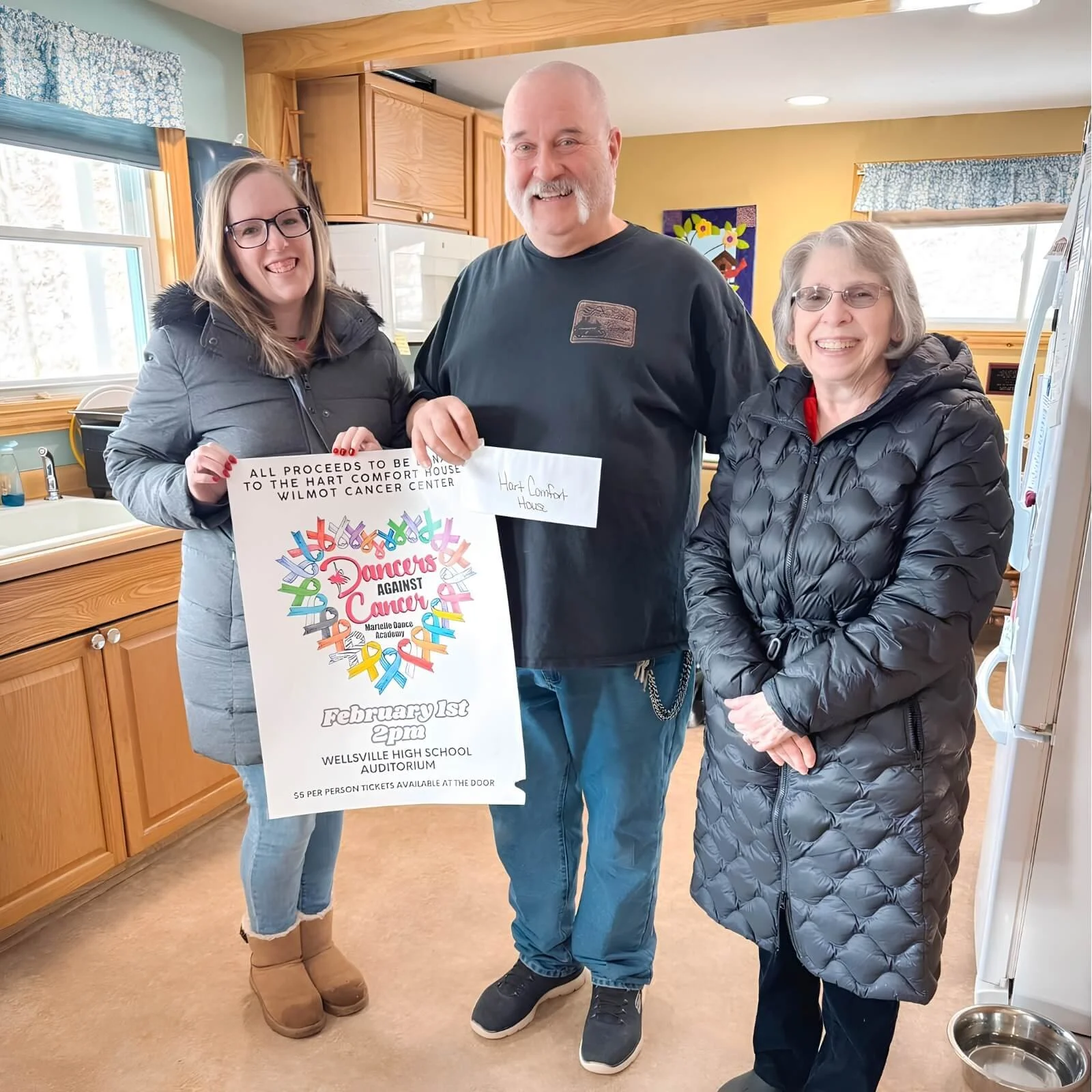Three women standing inside a kitchen, smiling and holding a poster for a fundraiser event. The poster is for Dancers Against Cancer, scheduled for February 1st at 2 pm at Wellsville High School Auditorium. 