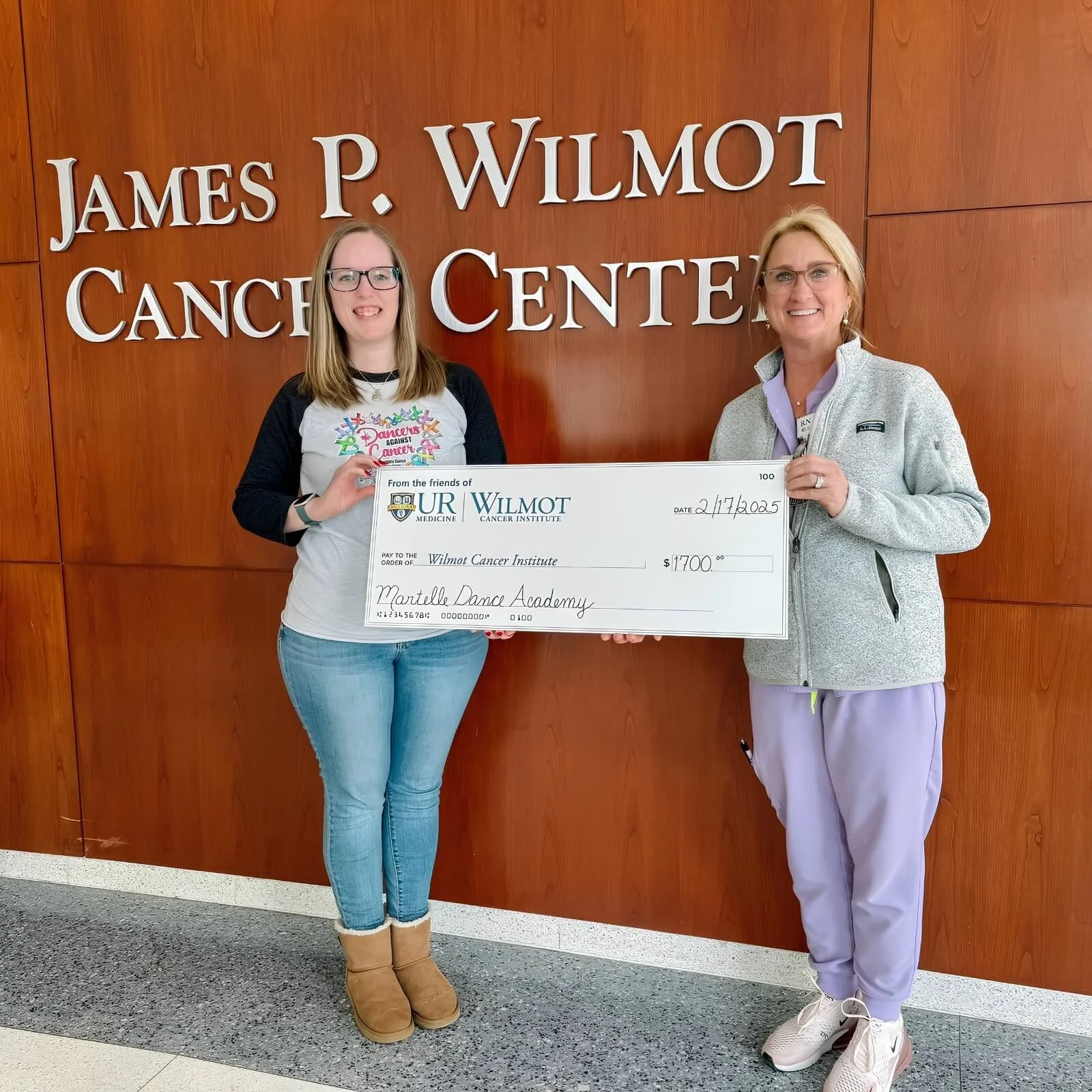Two women standing in front of a wooden wall with a sign that reads 'James P. Wilmot Cancer Center'. They are holding a large donation check made out to the Wilmot Cancer Institute for $1700, dated February 17, 2025. 