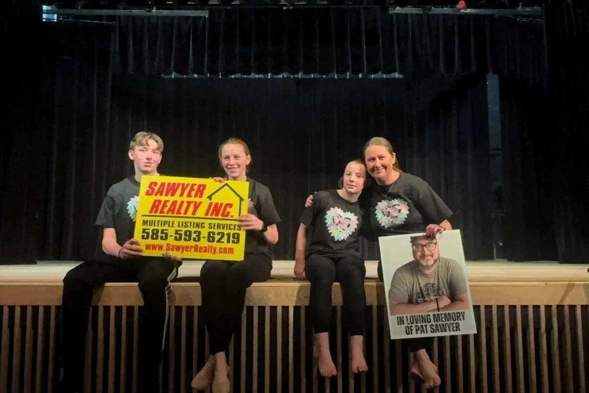 Four people, three females and one male, sit on a stage with a black curtain backdrop. They are holding signs and a photo in a memorial setting.