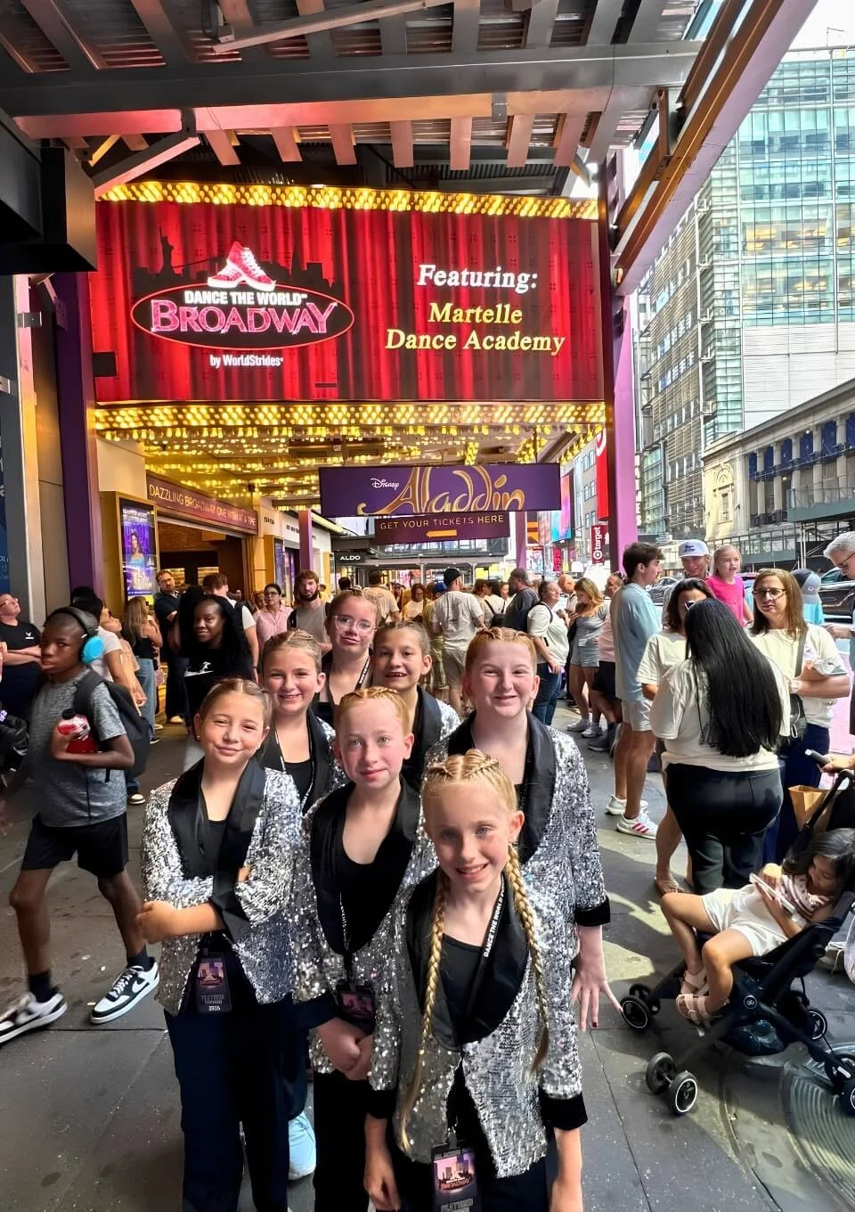 A group of young girls wearing matching silver sequin jackets and black tops posing together outside a Broadway theater on a busy city street, with crowds of people and tall buildings around.