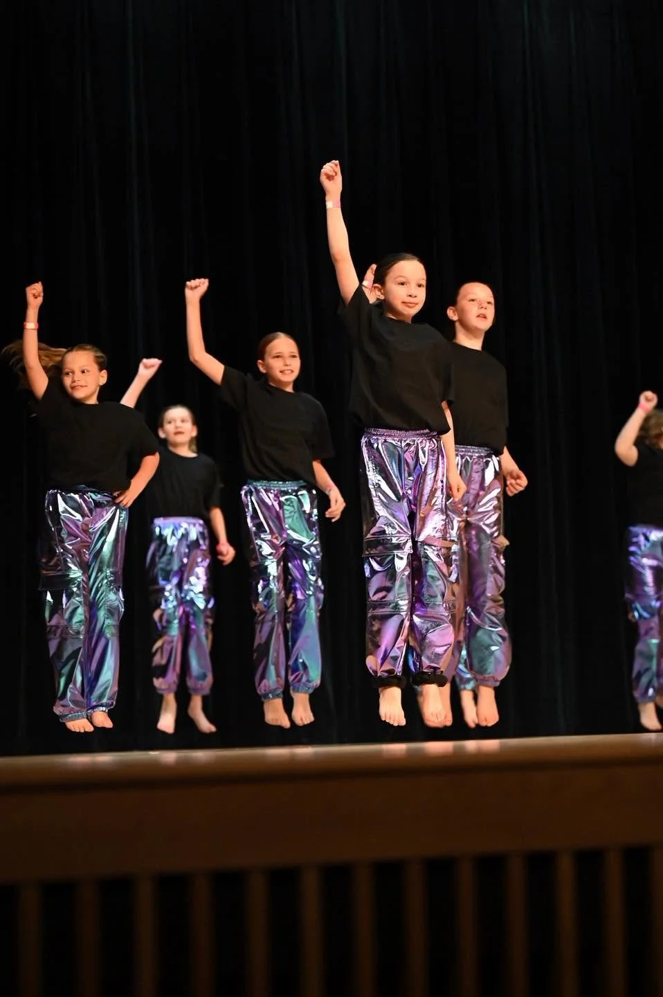 Young children performing a dance on stage, jumping with arms raised, dressed in black tops and shiny metallic purple pants, against a black curtain background.