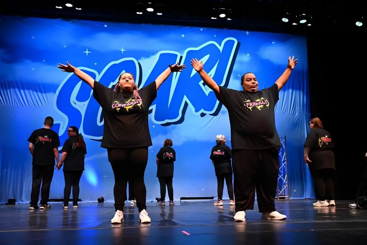 Group of performers on stage with arms raised, wearing matching black shirts with colorful text, during a performance at an event called 'SOAR'.