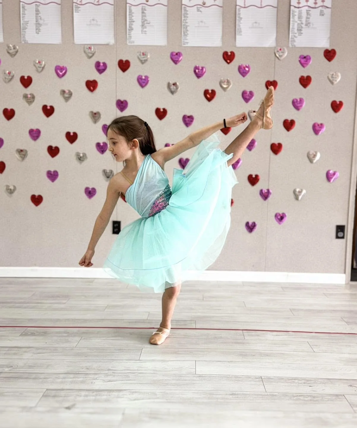 Young girl in a light blue ballet dress performing a dance move in a studio with heart-shaped decorations on the wall.