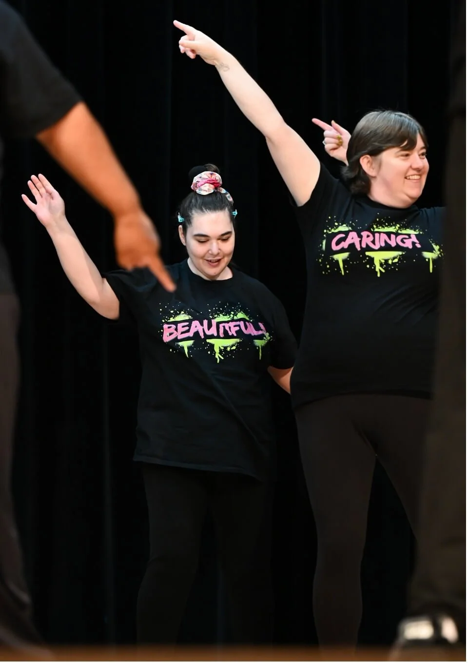 Two smiling women wearing black t-shirts with colorful text and designs, standing with arms raised, in front of a black curtain.