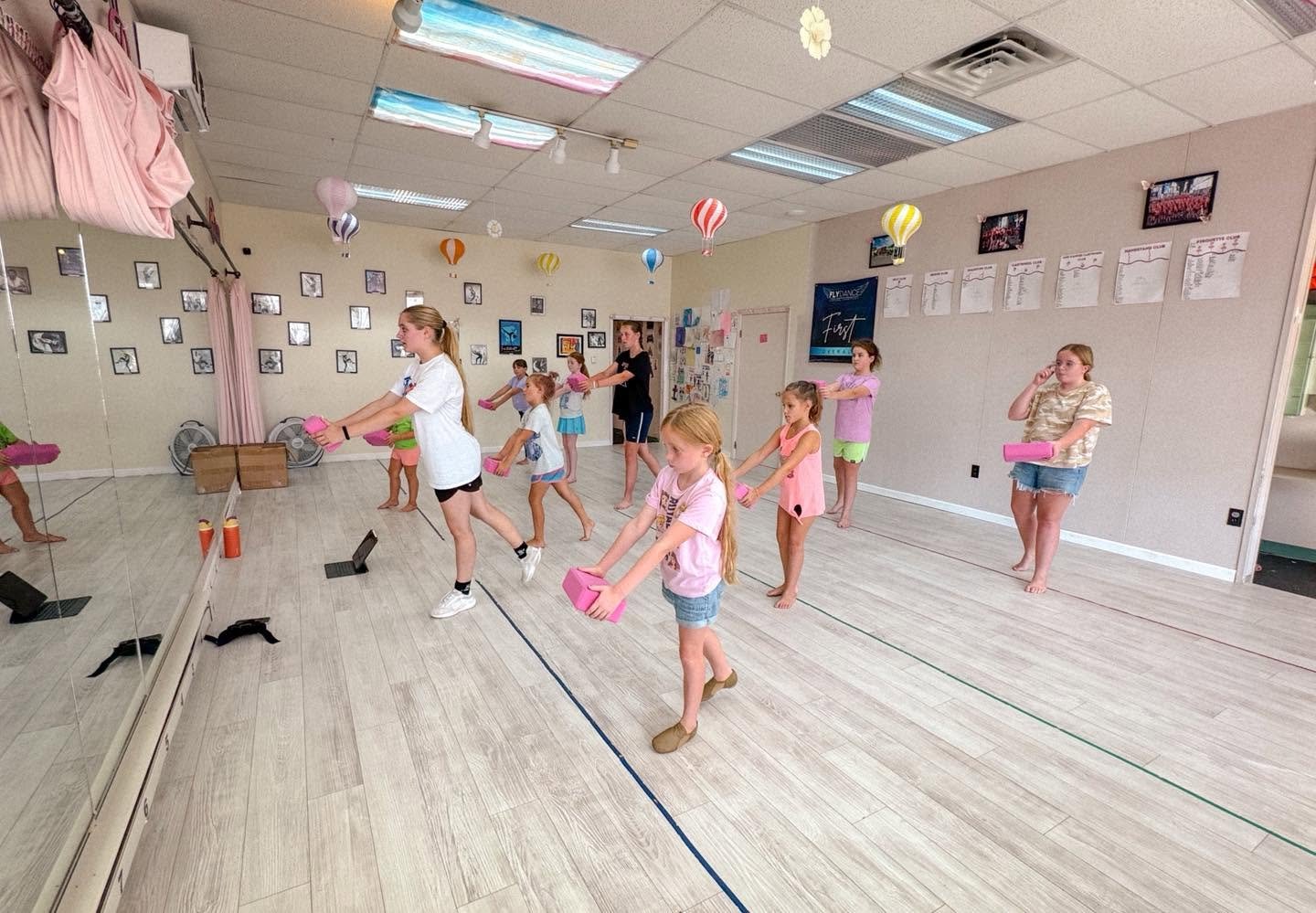 Children participating in a dance or exercise class in a studio, holding pink foam blocks, with a mirrored wall, framed photographs, and colorful balloons hanging from the ceiling.