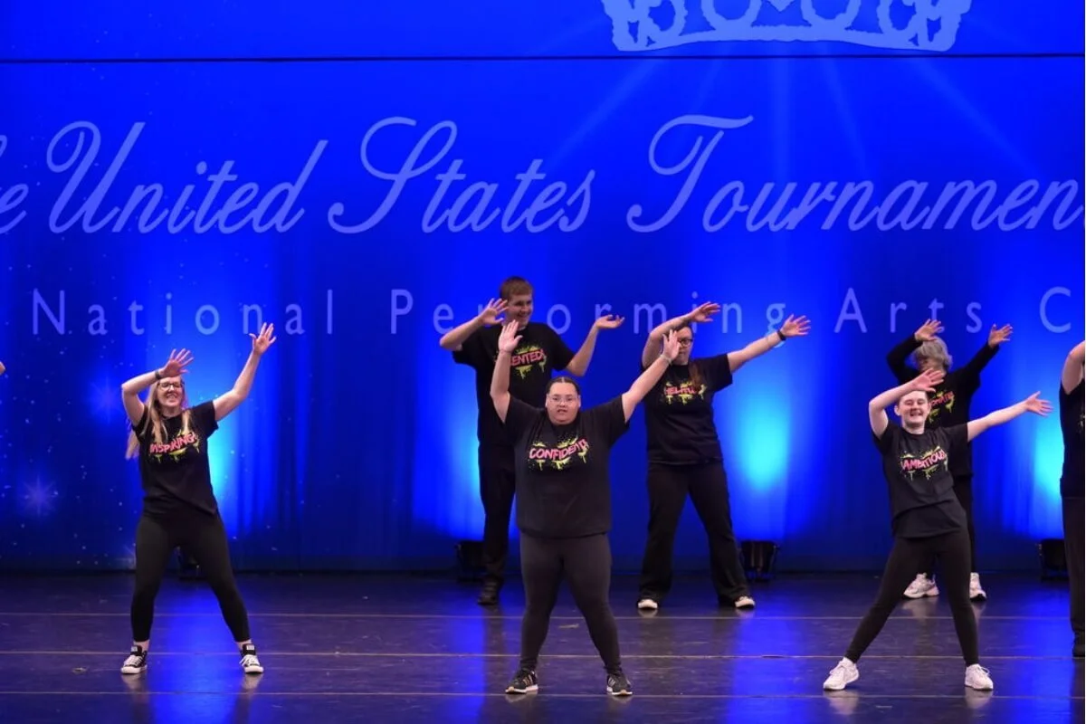 Group of performers on stage at the United States Tournamens of National Performance Arts with a blue backdrop, some raising their arms in a dance pose.