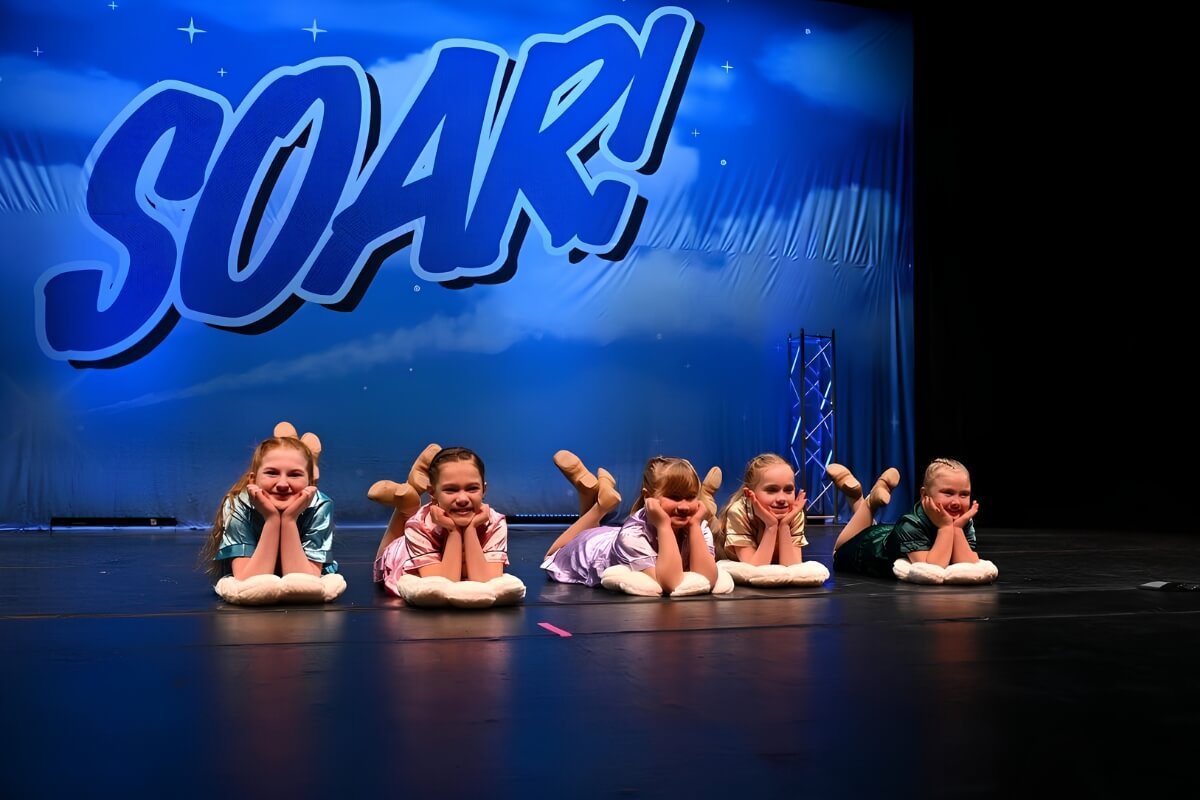 Five young girls in colorful pajamas lying on their stomachs on stage in front of a blue backdrop with the word 'SOAR!' and stars, performing a dance routine.