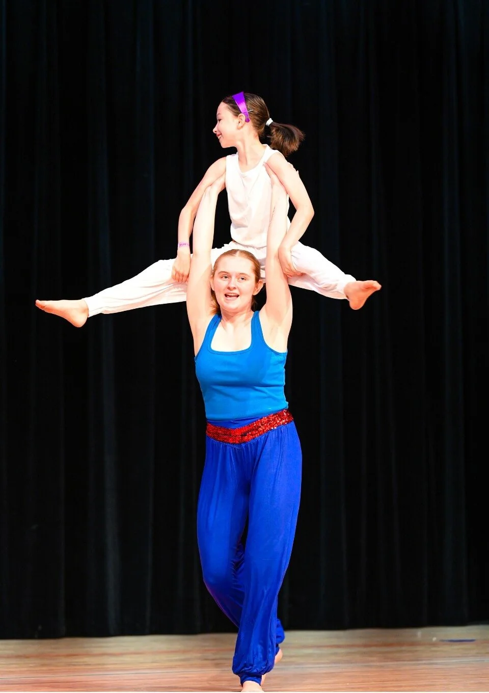 Two female acro dancers performing a balancing act on stage. One is standing and holding the other above her head with raised arms.