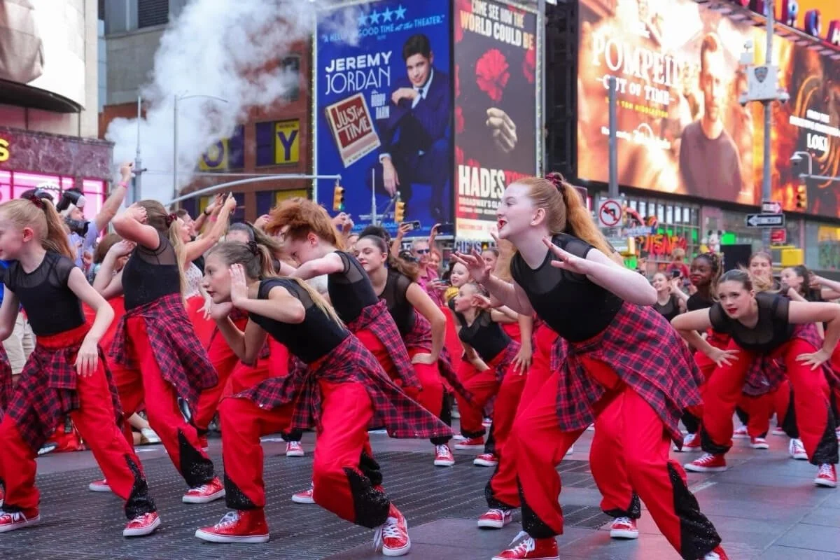 A group of children dancing in Times Square, New York City, with large billboards and advertisements in the background.