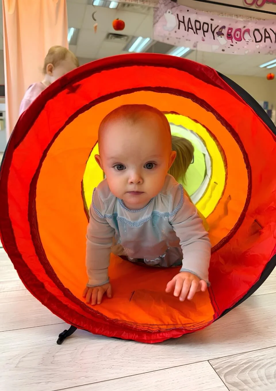 A baby crawling through a colorful, orange and yellow fabric tunnel with a serious expression, at the Baby & Me class offered at Martelle Dance Academy.