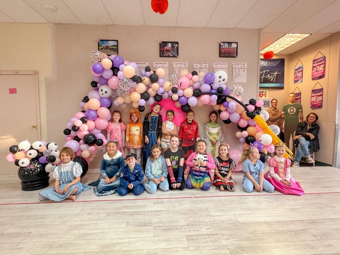Children in colorful costumes posing in front of a balloon arch decorated with pink, purple, black, and white balloons and paper lanterns inside a room, with some adults sitting on the side.