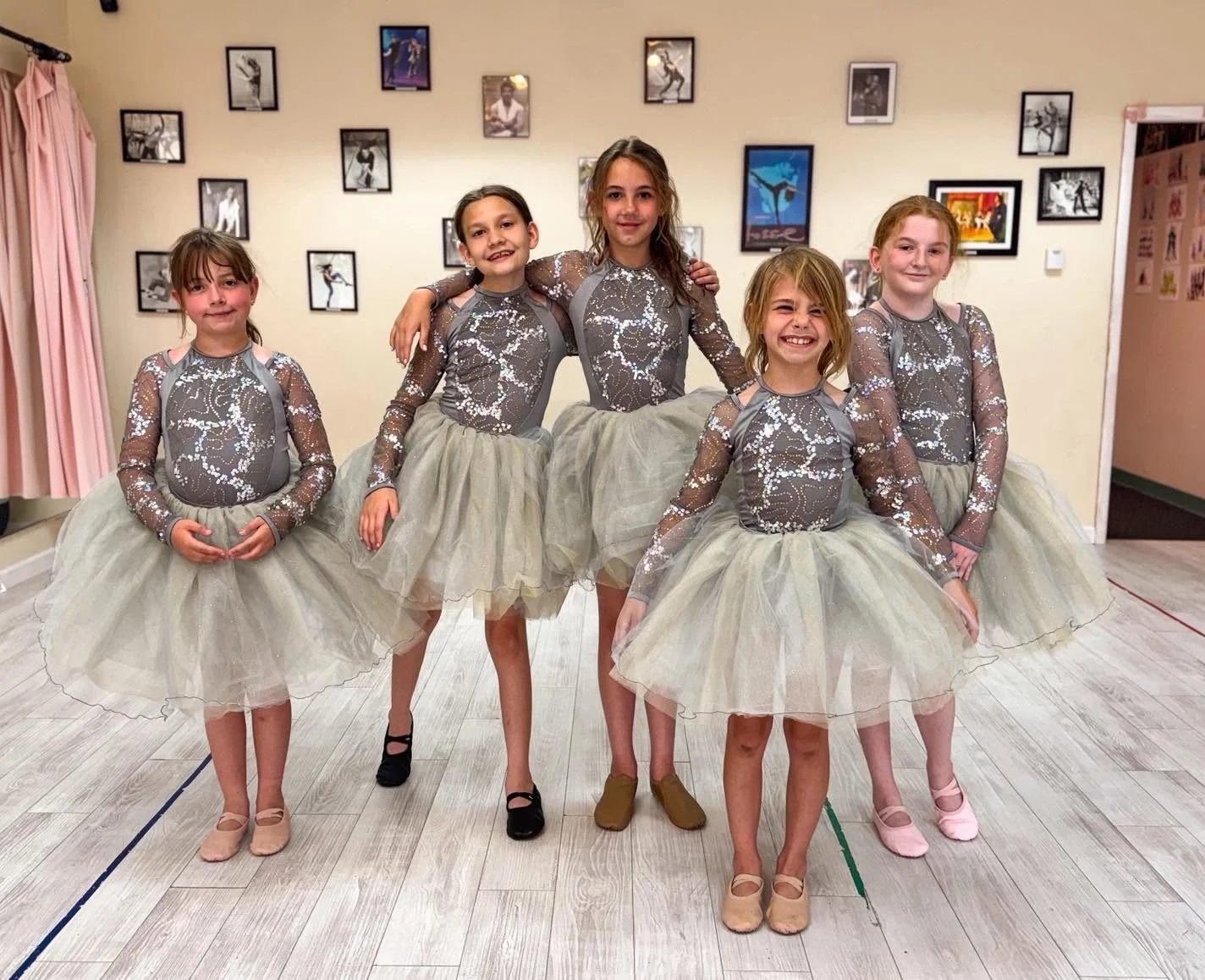 Five young girls in dance costumes with sparkly gray tops and beige tutus posing in a dance studio, standing close together with some girls smiling and others having neutral expressions.