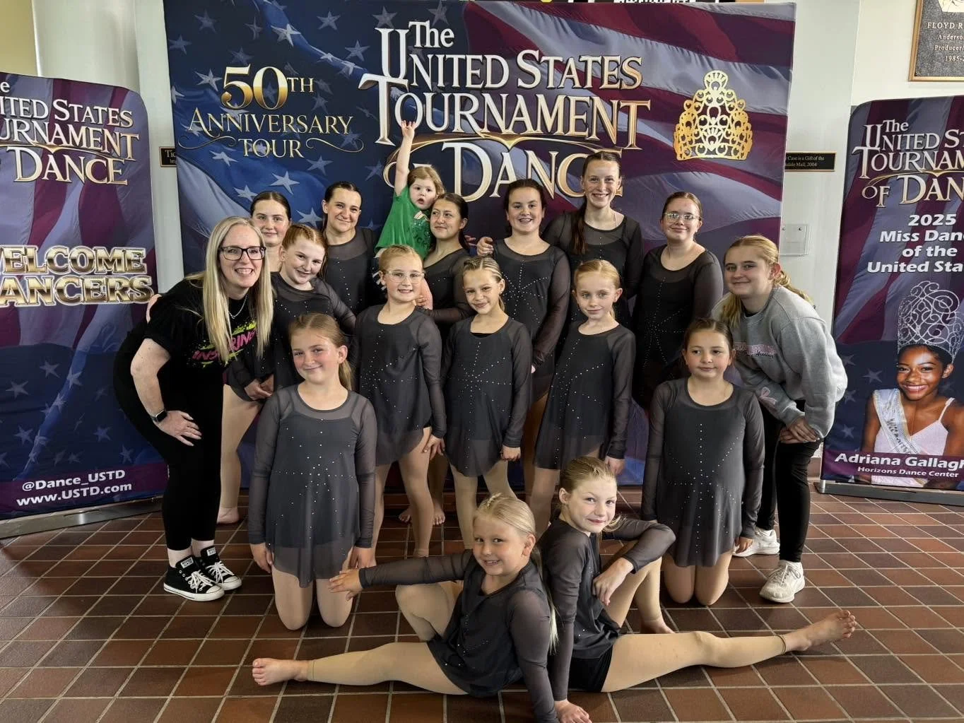 Group of young female dancers in black costumes with rhinestone details, posing for a photo at the United States Tournament of Dance event, with banners in the background celebrating the 50th anniversary and welcoming dancers.
