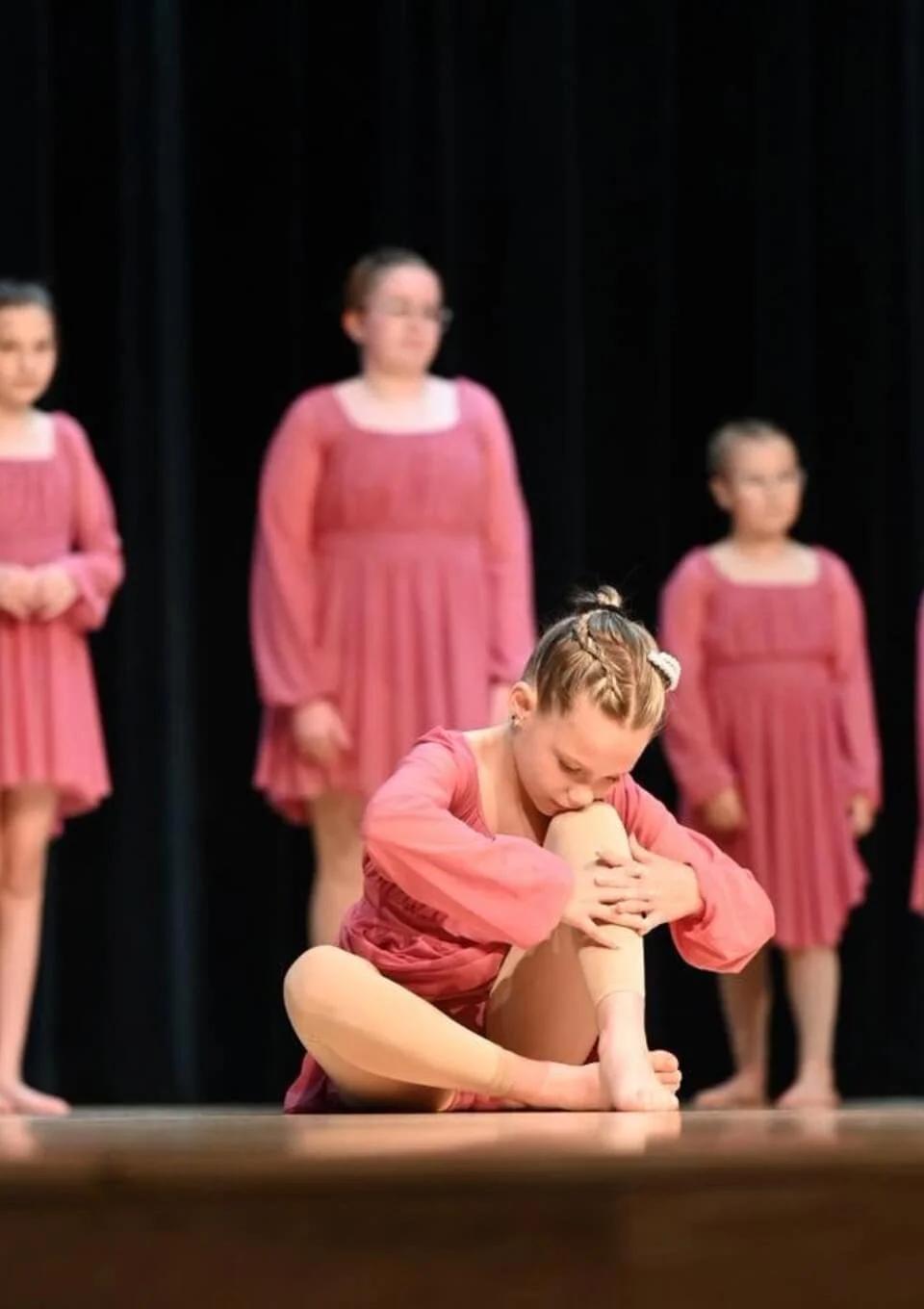 A young girl in a pink dress performing a dance move on stage, holding her leg with her hands, while other girls in similar dresses stand behind her watching.