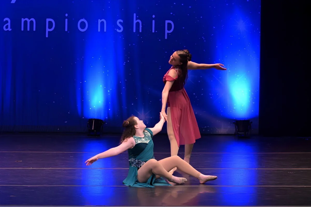 Two young ballet dancers perform on stage with a blue starry background. One dancer is sitting on the floor with one leg extended and holding the hand of the other dancer, who is standing with arms outstretched.