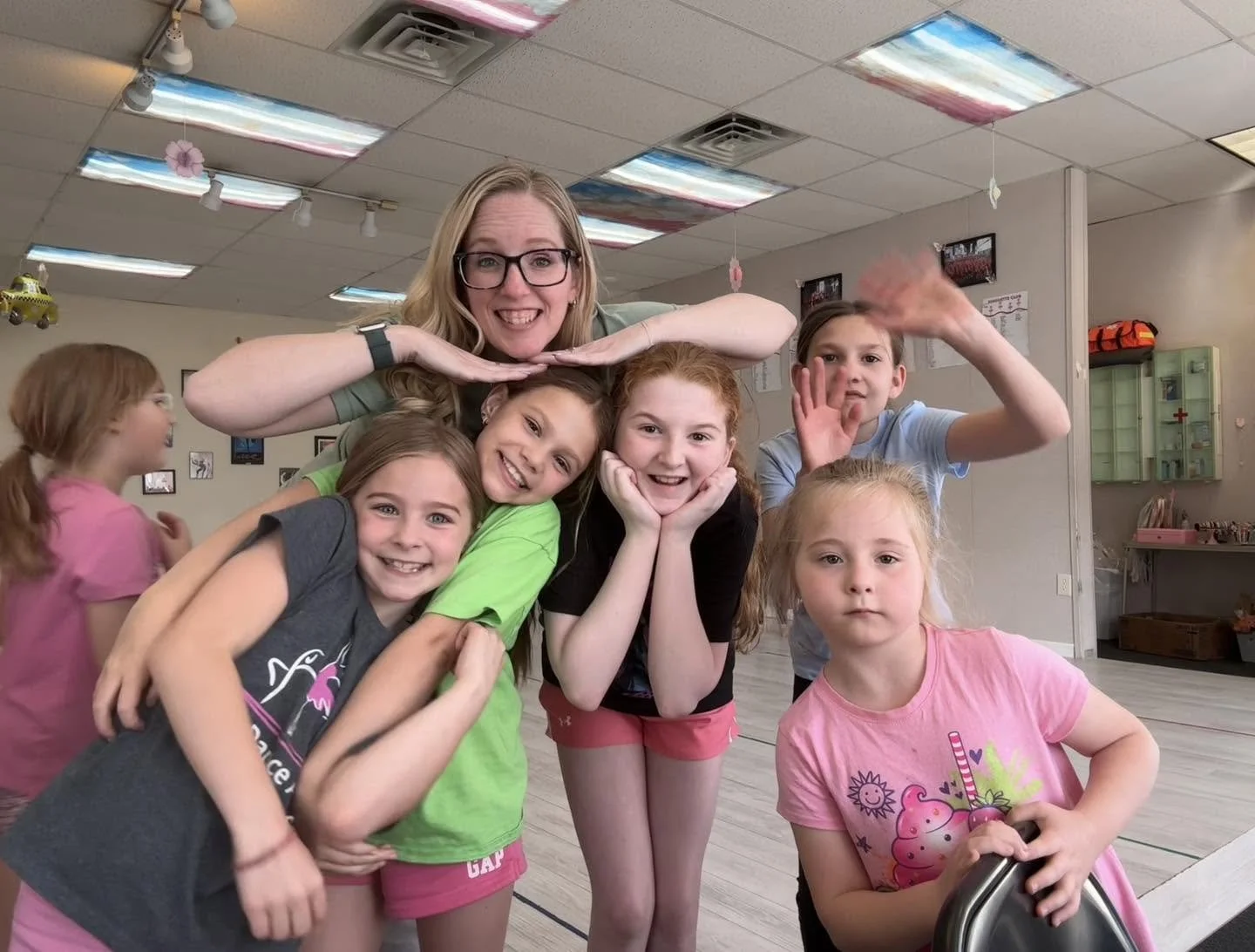 A group of six smiling young girls, with one woman, posing playfully inside a dance studiowith colorful ceiling tiles. The women and girls are making fun poses, with some leaning on each other and others waving.