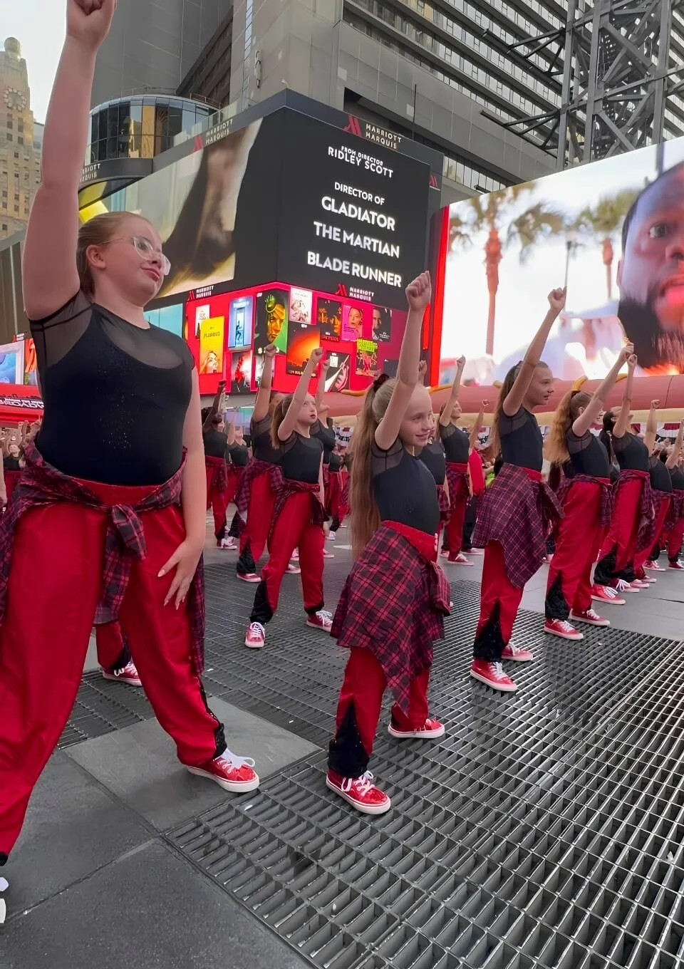 Group of young girls dressed in black tops, red pants, and red sneakers with plaid shirts tied around their waists, performing a choreographed dance or movement routine in Times Square, New York City.