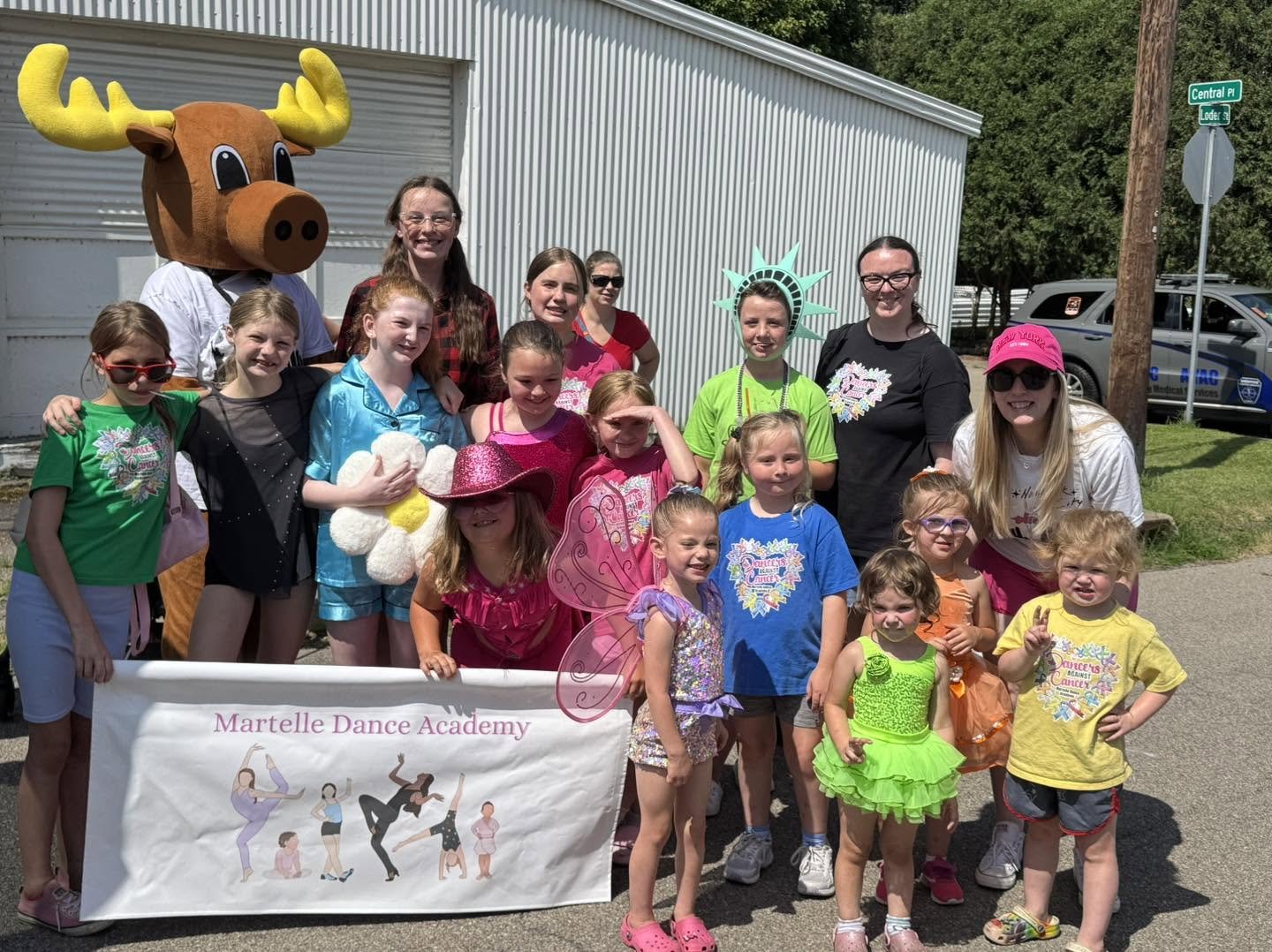 Group of children and adults posing outdoors with two costumed characters, one dressed as a moose and the other as the Statue of Liberty, holding a banner that reads "Martelle Dance Academy."