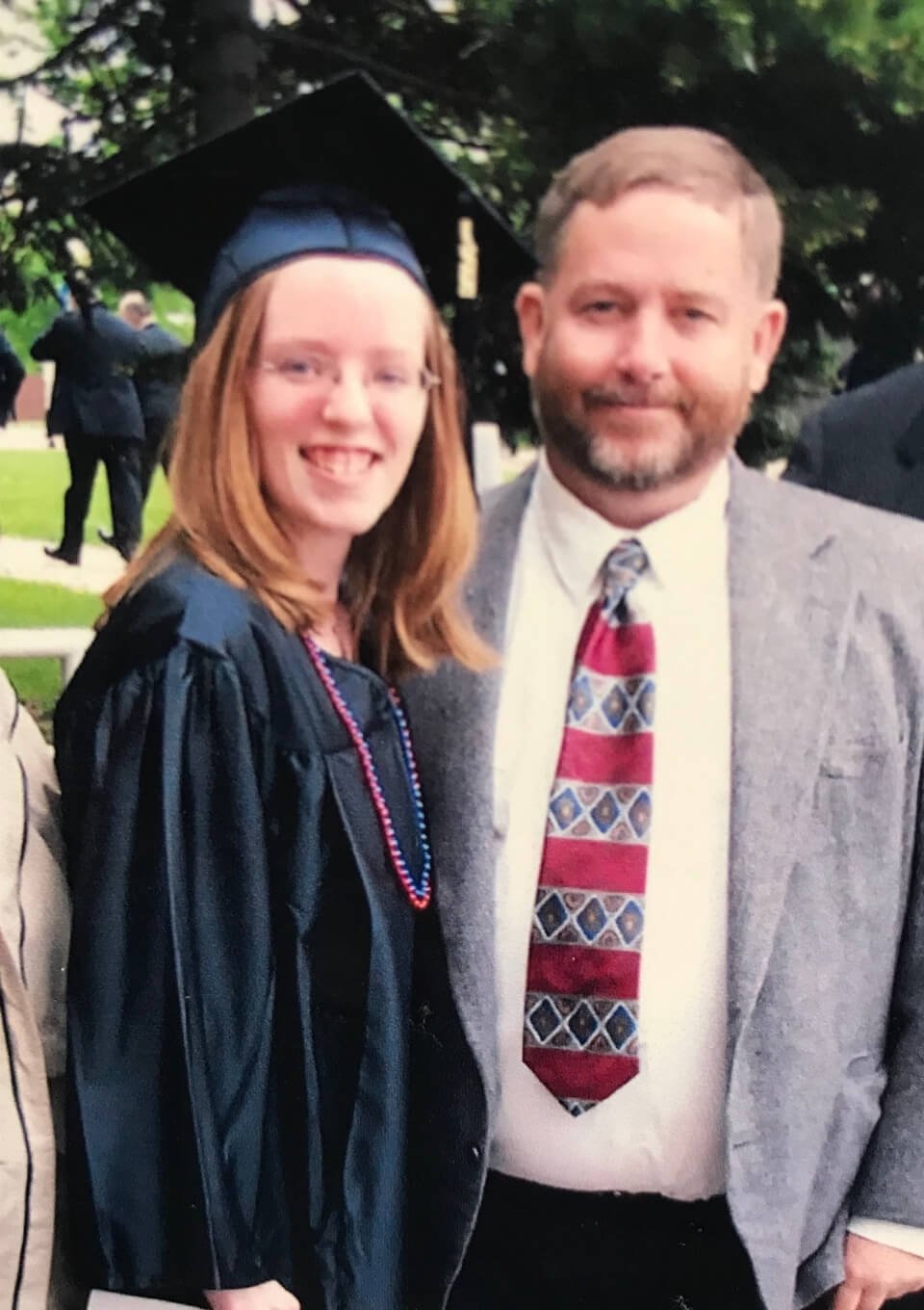 A woman in a graduation cap and gown smiling with a man in a gray blazer and patterned tie, outdoors with trees in the background.