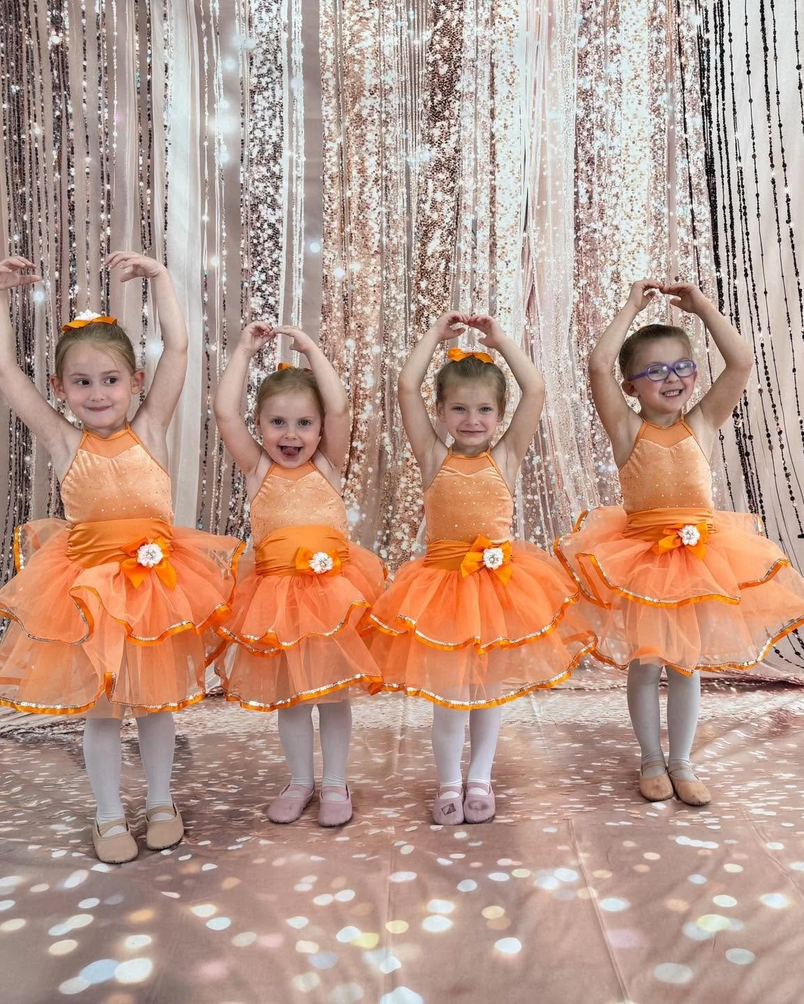 Four young girls dressed in matching orange ballet costumes, standing in front of a sparkly, sequin-covered curtain, posing with their arms raised.