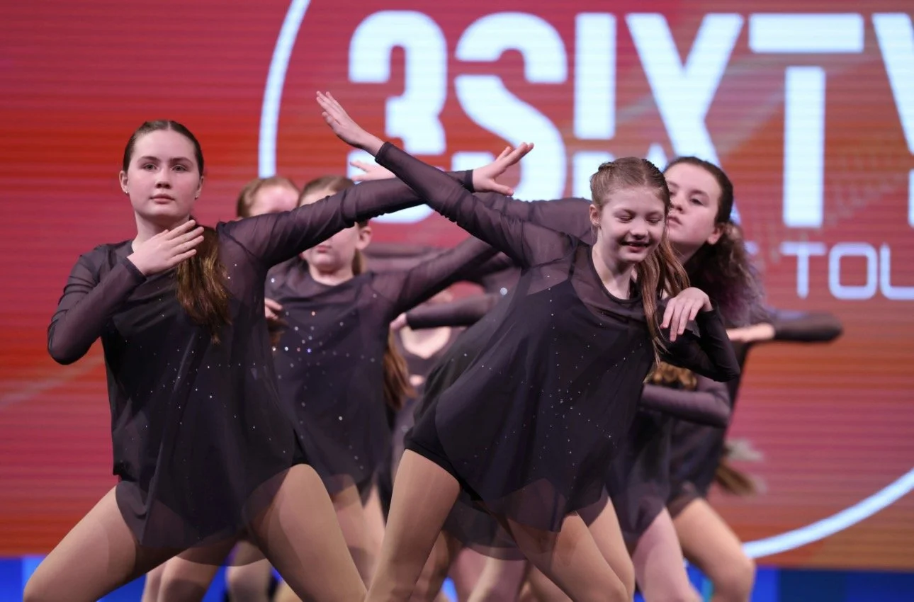 A group of young girls performing a dance on stage in black outfits with sparkles, with a red background featuring a large logo.