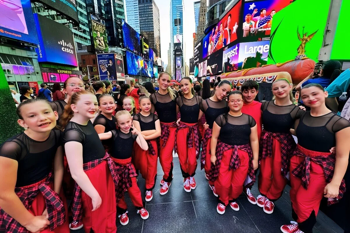 Young girls and women in matching dance costumes with black tops, red pants, and plaid flannel shirts tied around their waists, posing together in Times Square, New York City, with bright billboards and crowds in the background.