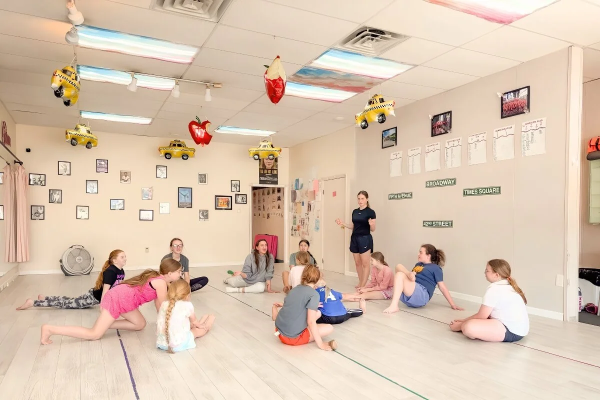 A group of children sitting on the floor in a dance studio, listening to an instructor who is standing and speaking. The room is decorated with hanging yellow taxi and strawberry balloons, and framed photos and signs on the walls.