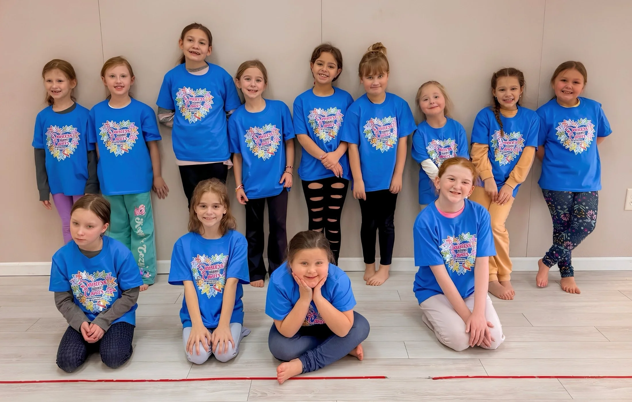 Group of young girls wearing matching blue T-shirts posing for a photo indoors, smiling and sitting on the floor.