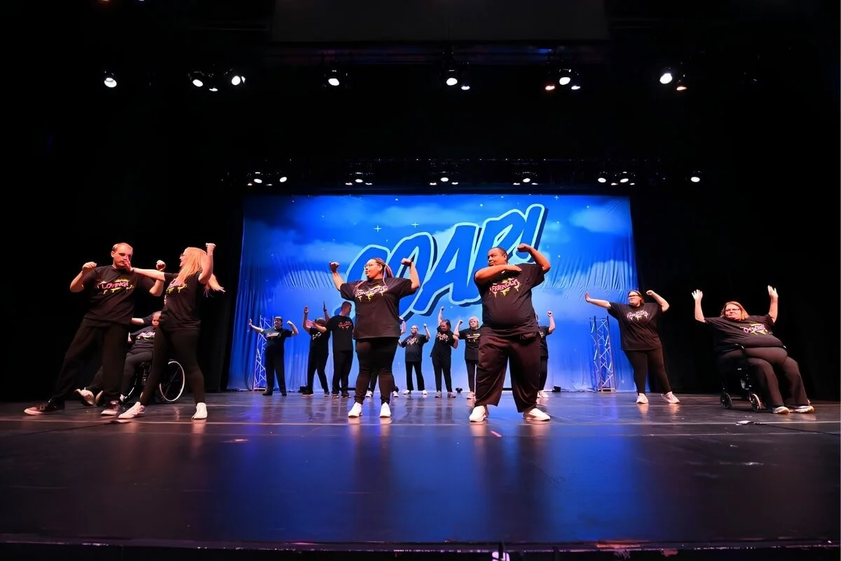 Group of performers dancing on stage during a live show, with stage lights and a large screen displaying the word 'SOAR' in the background.
