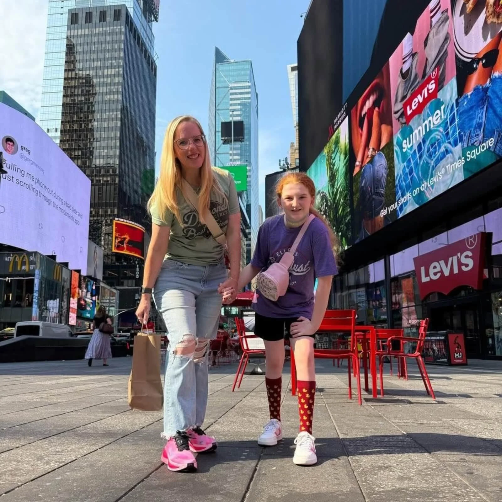 A woman and a girl holding hands in Times Square, New York City, with tall buildings and digital billboards in the background.