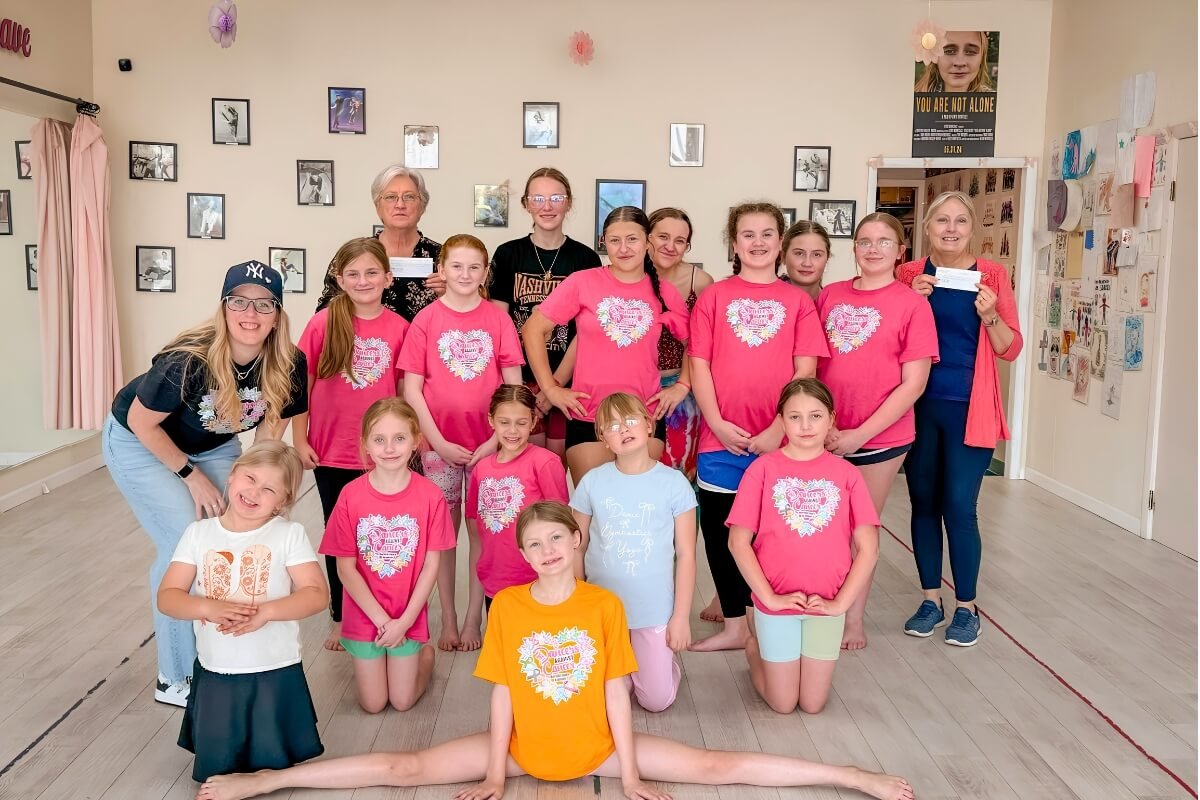 Group of children and adults posing indoors, some children wearing pink T-shirts with a white heart design, inside a room decorated with framed pictures on the wall and pink curtains.