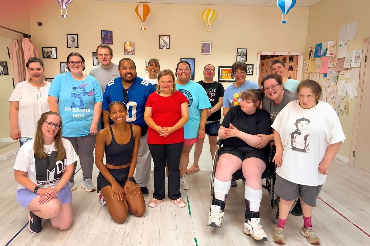 Group of diverse smiling people, including young, middle-aged, and one person in a wheelchair, posing together in a room decorated with hanging paper lanterns and wall art.