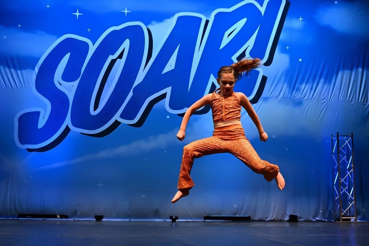 A young girl in an orange and brown dance costume jumps on stage during a dance performance with a large blue backdrop reading "SOAR" in the background.