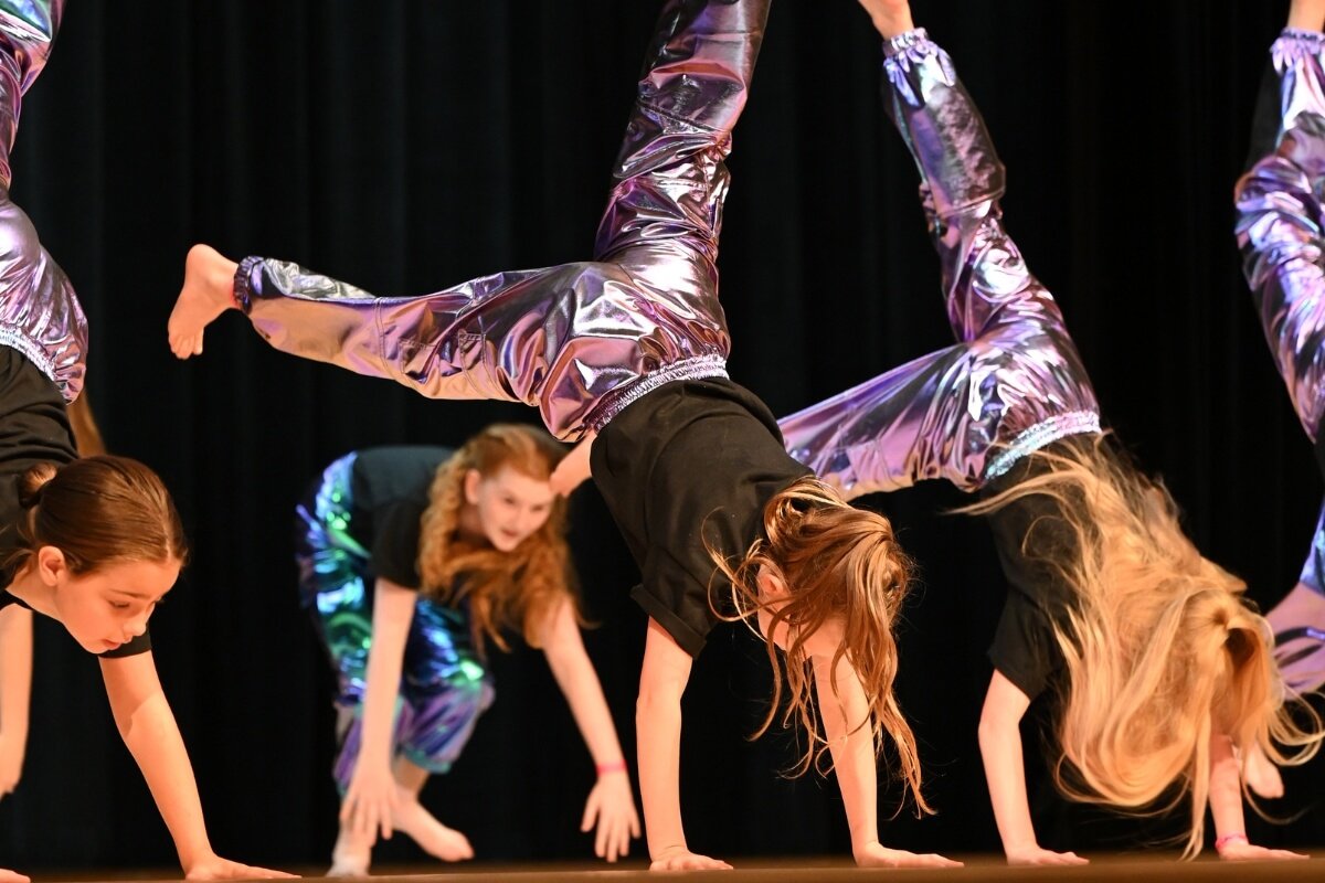 Group of young girls performing a dance routine on stage, all in handstand positions with some girls having their legs extended and bent, wearing black tops and shiny purple pants.