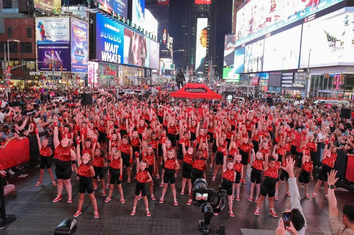 A large group of young dancers in red and black outfits performing in Times Square, New York City, at night with bright advertisements and billboards in the background.