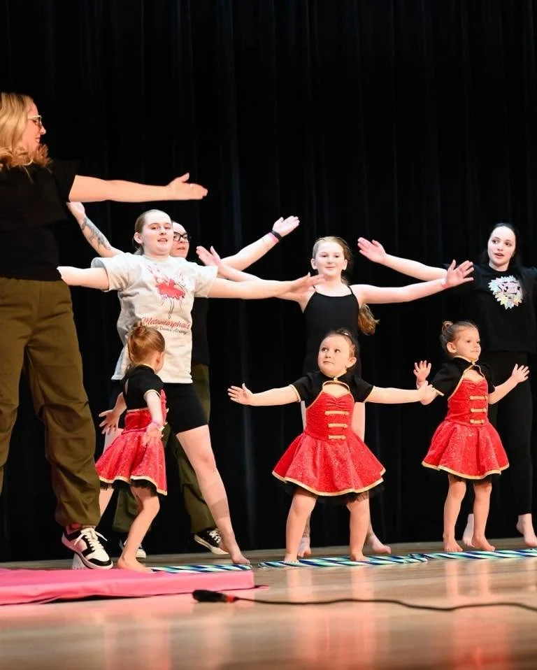Young girls performing a dance on stage with black curtains background, wearing red and black costumes, with some adults guiding them.